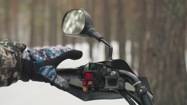 Gloved hand on handlebars in snow, camouflage jacket testing throttle and switches, mirror reflecting pine forest, closeup grip and ignition, focused prep for extreme offroad ride