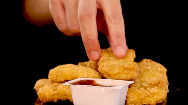 Dipping golden chicken nugget into barbecue sauce container over black background in slow motion cinematic video clip.