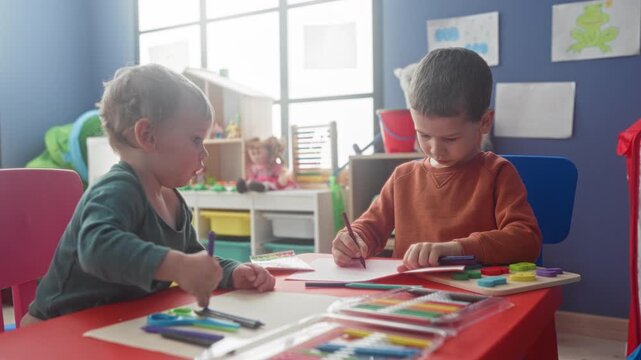 Boys drawing at a small table with pencils and crayons, toddler reaching with hand while older brother colors in a preschool building; concentration sibling bonding.
