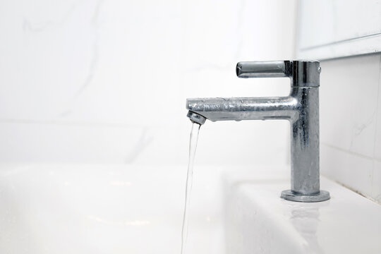 Close-up of a modern chrome-plated bathroom faucet on a white marble vanity, with a clear stream of water running from its spout. Perfect for concepts of freshness, cleanliness, and water conservation