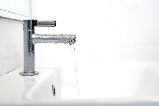 Close-up of a modern chrome-plated bathroom faucet on a white marble vanity, with a clear stream of water running from its spout. Perfect for concepts of freshness, cleanliness, and water conservation