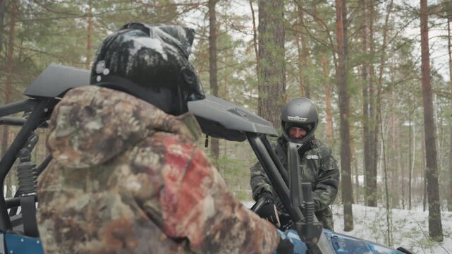 Two riders approach atv in snowy forest before extreme ride, one prepares to board while other checks engine and secures gear helmeted figures exchange signals, fastening harness and goggles, visible