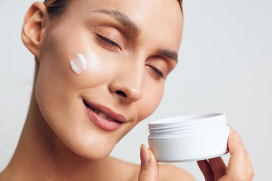 Woman skincare cream beauty jar face closeup cosmetic moisturizing moisturizer, young woman applies soft moisturizer on smooth skin, holding white jar, eyes closed, calm spa beauty routine on neutral