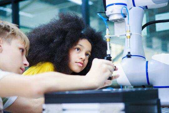 Young Black girl and boy intently collaborate with a robotic arm in a brightly lit classroom, exploring STEM learning and technological innovation with a blue robotic arm, focused on future robotics a