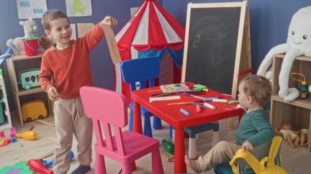 Boy holds crayon drawing up to camera showing fingers and paper in a playroom inside building; playful curiosity.