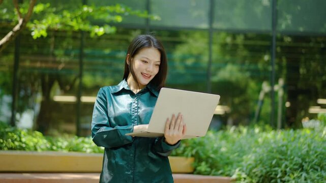 Happy young Asian business woman working on her laptop in a green outdoor park near modern office building. She enjoys remote work and digital lifestyle while smiling at her computer screen, Freelance