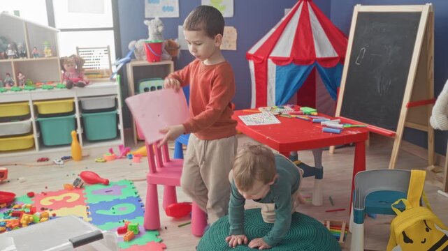 Boy holds pink paper with tongue out while younger brother sits by scattered toys inside building; childhood curiosity.