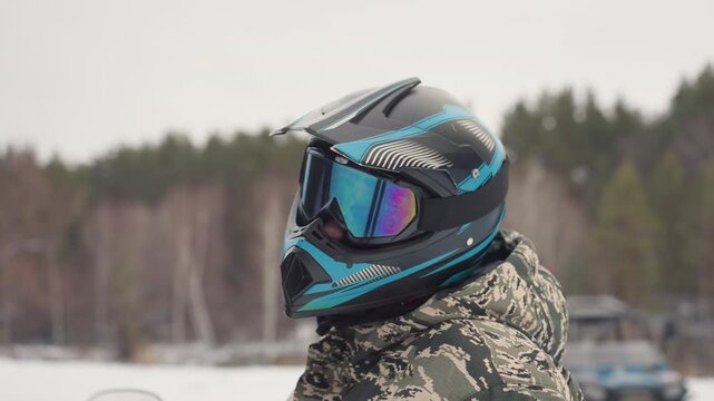 Atv rider with reflective helmet, camo jacket, snowy forest background, mirrored goggles catching overcast light, steady preride posture, checking visor and straps, calm anticipation, soft cold
