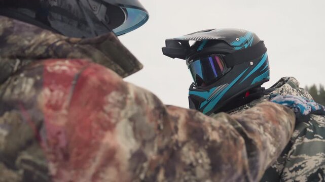 Closeup winter helmet check between riders, camo jackets and reflective goggles, instructor secures strap while snowmobile waits at edge of pine forest, tense supportive camaraderie before extreme