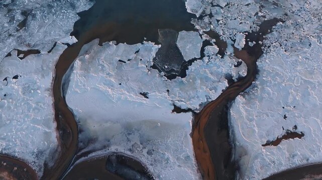 Stunning aerial drone footage of frozen river channels cutting through ice and snow in South Iceland. Abstract patterns of dark water, rust-colored sediment, and white ice create a dramatic winter lan