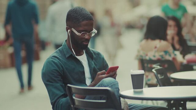Man enjoys coffee, Man relaxed with phone and espresso, Man lounges at cafe with glasses and earbuds