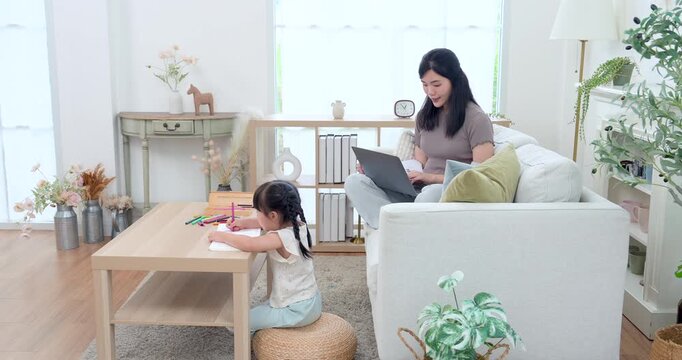 Asian single mother working on laptop from sofa while preschool daughter draws at coffee table, wide home learning scene with parent supervision, early education, childcare support, warm family bond