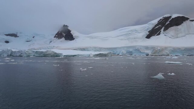 Cinematic POV shot moving slowly past towering blue-tinted ice walls and floating brash ice at the base of snow-capped mountains in the Antarctic Peninsula