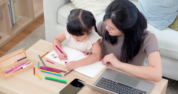 Overhead view of Asian mom coaching preschool daughter while girl draws simple picture with pink marker beside laptop, phone, notebook, and crayon, reflecting guidance, development, and study time.