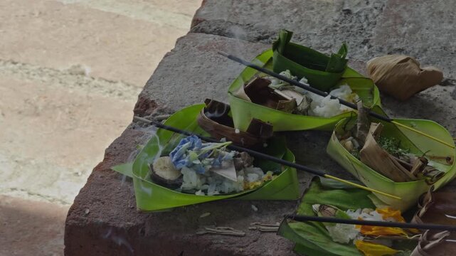 A close up of Balinese canang offering placed on the ground, made from coconut leaves filled with flowers and burning incense. Represents daily Hindu prayer rituals and sacred cultural tradition.