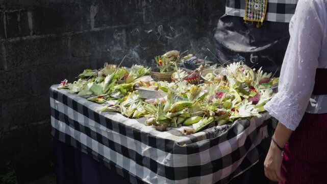 A close up of Balinese canang offering placed on a table beneath a temple shrine, made from coconut leaves with flowers and burning incense. Captures sacred ritual, culture, and spiritual devotion.