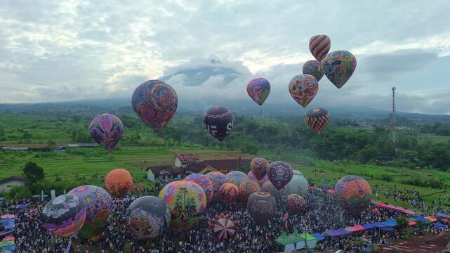 Colorful hot air balloons fill the sky during a lively festival. Aerial view of crowd, tents, and scenic countryside landscape. Wonosobo balloon festival, Indonesia.