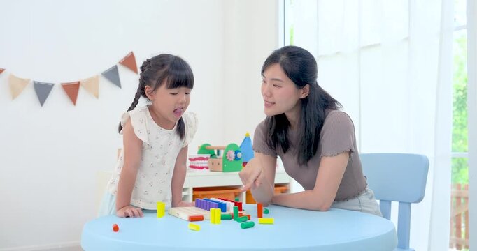 Asian single mother pointing while preschool daughter arranges colorful rods on board game at table, interactive home learning play supporting logic, concentration, coordination, child confidence