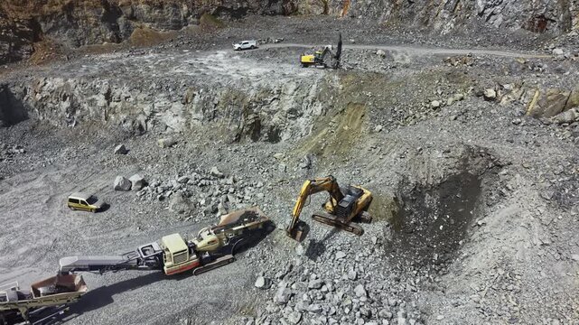 Heavy machinery is seen working in a granite quarry. The equipment is involved in rock extraction and processing. Trucks are transporting materials within the site.