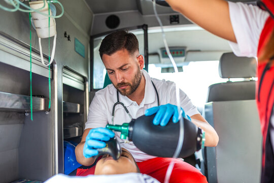 Male paramedic using a manual resuscitator bag to help a patient breathe inside an ambulance. Emergency medical technician performing life-saving CPR procedure. Healthcare and medicine concept.