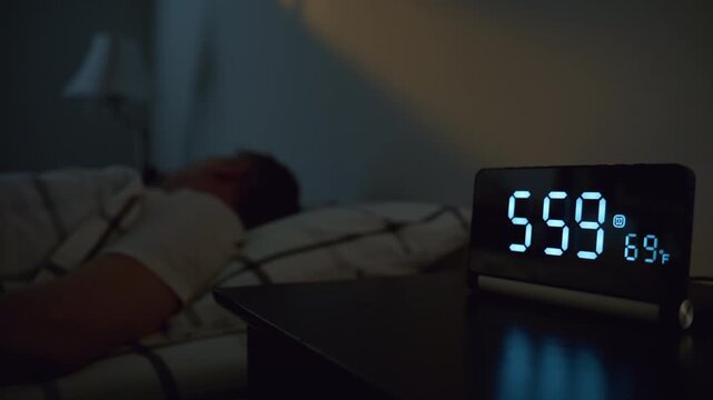 Man turning off alarm clock at 6.00 in bedroom. Early morning wake up with digital clock beside bed, hand reaching to stop alarm