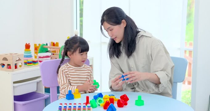 Asian single mother smiles beside preschool daughter arranging colorful shape blocks at home table, encouraging fine motor skill, concentration, and warm parent child development through play.