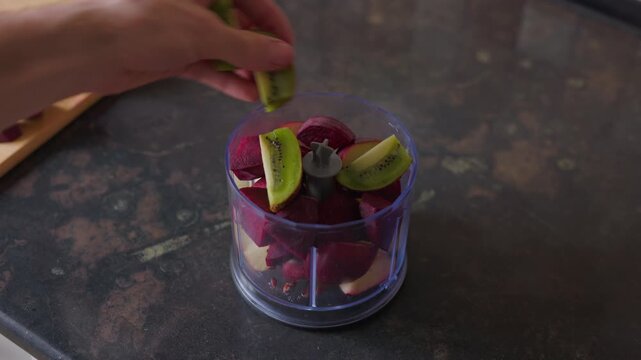 Static tripod shot of a person adding kiwi slices to a blender with beets and apples
