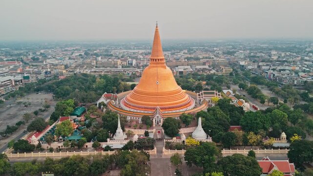 High angle drone perspective pulls away from huge golden stupa rising above urban center while majestic Buddhist monument dominates hazy gray city skyline in Thailand during serene beautiful morning