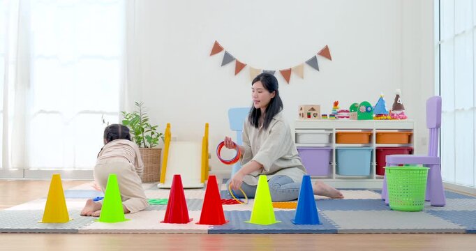 Asian single mother kneels in playroom and coaches preschool daughter through ring toss game, supporting balance, motor skill, and confidence as child prepares colorful hoop for active learning play.