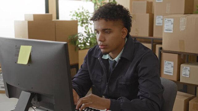 Young hispanic man at computer surrounded by parcel boxes, typing and finger to lips for silence in shipping building; concentration.