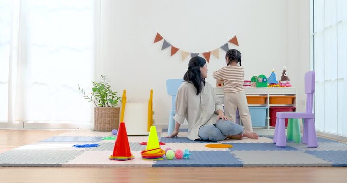 Asian single mother turns toward preschool daughter reaching toy shelf after movement task, supporting independence, listening skill, transition routine, and active learning in bright home playroom.