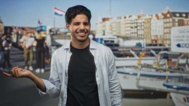 Man holds hand out and smiles, standing on a street by canal boats and rows of buildings; contentment easygoing.