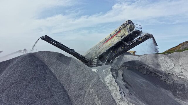 Granite is being processed by a machine at a quarry. Stone is being crushed and moved onto a large pile. Dust is rising from the activity in the background.