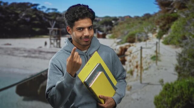 Man holding yellow folders and pointing finger toward camera while smiling on a sunny beach near rocks and sand; playful invitation.