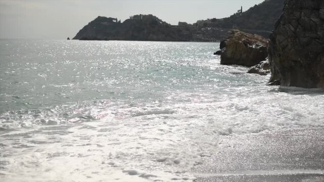 Gentle waves break on a pebble beach, with rugged cliffs and distant hills under a bright sky