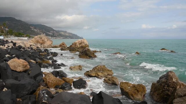 Dynamic ocean waves surge and break against a rugged, rocky coastline dotted with large boulders and a distant Mediterranean village nestled on a hillside