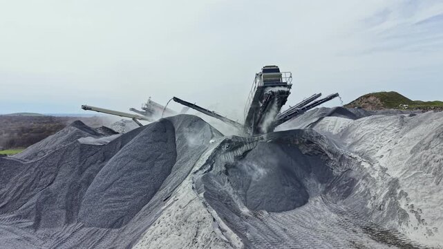 Workers are using large machinery to process granite in the quarry. Heavy equipment moves large amounts of rock and creates dust in an outdoor setting with hills in the background.