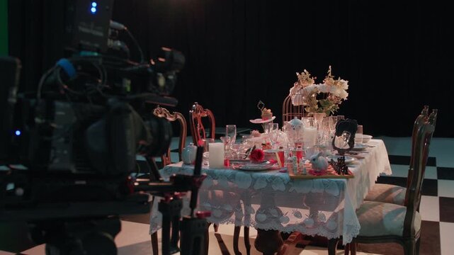 Behind-the-scenes shot of a stylized tea party table set on a film studio stage, featuring vintage tableware, flowers, candles, and whimsical props including a chessboard and white rabbit figurine.
