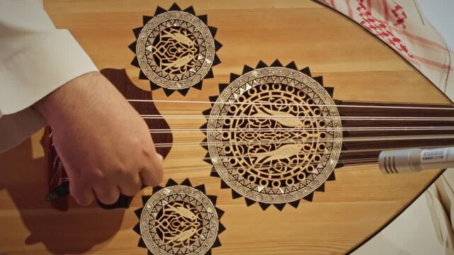 A detailed close-up of a musician&rsquo;s hand plucking the strings of a traditional
Saudi Arabian oud. The wooden instrument features intricate decorative
rosettes and craftsmanship.