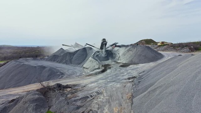Large piles of granite rocks fill the landscape. Heavy machinery moves stone in a granite quarry. Dust rises into the air as work continues under a cloudy sky.