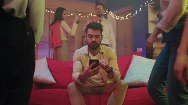 Man observes messages during lively social event, Individual glances at device as friends converse happily nearby