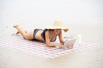 Southeast Asian woman relaxes on a sandy beach with a blue and white checkered blanket, enjoying a...