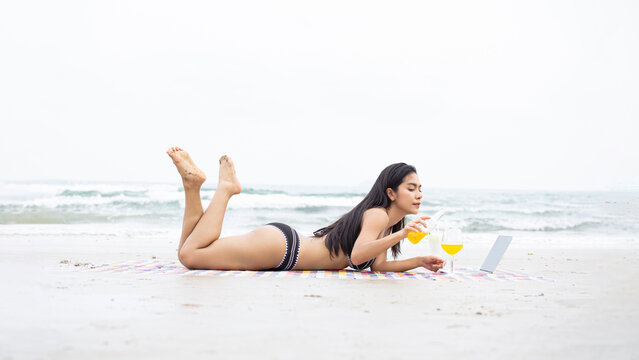 Southeast Asian woman relaxes on a sun-drenched beach, enjoying a chilled juice and creating a relaxed mood while wearing a bikini in this beachfront setting, featuring a blue ocean and sandy shore.