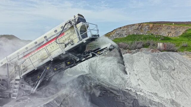 A machine works at a granite quarry, breaking large rocks into smaller pieces. Dust rises as the process continues under clear skies in a remote location.