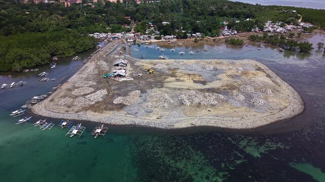 Backward drone flight revealing stone covered reclamation mound beside coastal settlement and mangroves near Bancao-Bancao in Puerto Princesa on Palawan island Philippines under cumulus sky