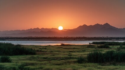 Obraz premium A vibrant sunset casts a warm glow over a tranquil lake, framed by lush green reeds and a distant mountain range, creating a serene and picturesque landscape.