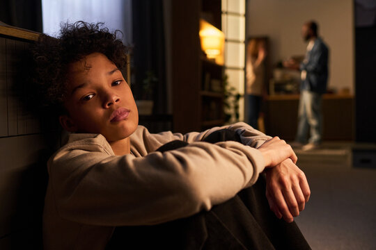 Portrait of teenage boy sitting alone and looking at camera while parents arguing in background, expressing family stress and emotional conflict. Useful for parenting, divorce