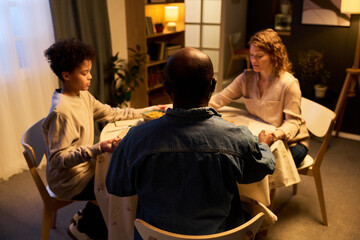 Obraz premium Multiethnic family praying before dinner at home, joining hands around table and practicing faith ritual. Suitable for parenting, family values, religion, togetherness marketing