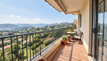 Obraz premium Sunlit balcony overlooks green hills and distant mountains. Metal railing borders tiled patio with potted plants. Small table and chairs invite relaxation under bright sky
