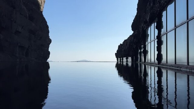 Still water reflects cliff walls and a row of glass-fronted structures along Tianmen Mountain, with distant mountains visible across the horizon near the 999 steps area of Stairways to haven.